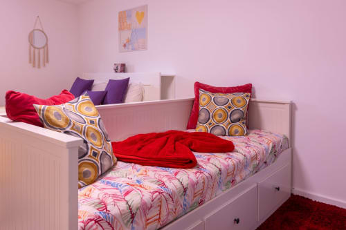 Quarto infantil colorido com paredes rosa, cama branca, almofadas com padrões geométricos e decoração de parede no apartamento EcoHost.