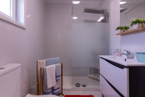 Banheiro moderno com chuveiro espaçoso, lavatório, sanitário, toalheiro em madeira, plantas em vasos e iluminação clara em Perafita