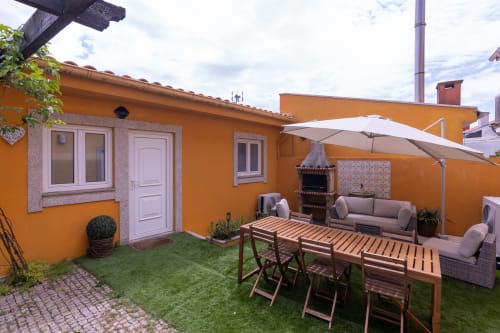 Casa amarela brilhante com terraço, mesa de jantar em madeira, área de estar, guarda-sol branco e jardim com grama sintética na EcoHost