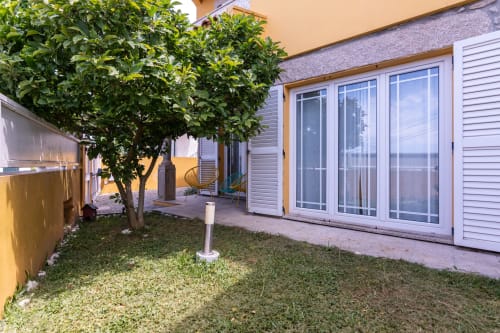 Pátio do jardim da Casa Amarela de Perafita com grama, árvore verde e portas de vidro que abrem para área de estar externa