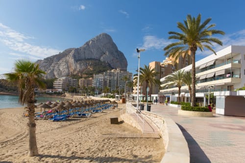 Playa de Calpe con tumbonas azules, palmeras y la formación rocosa del Peñol d'Ifach dominando el Hotel Galeón