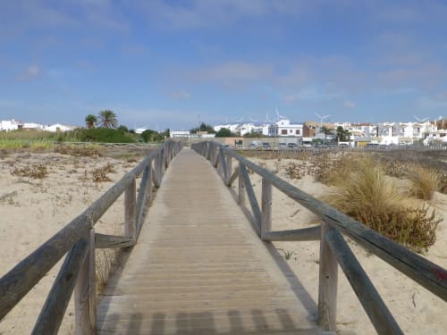 Pasarela de madera cruzando playa arenosa hacia Barrosa Home en Cádiz, España, con edificios blancos y palmeras