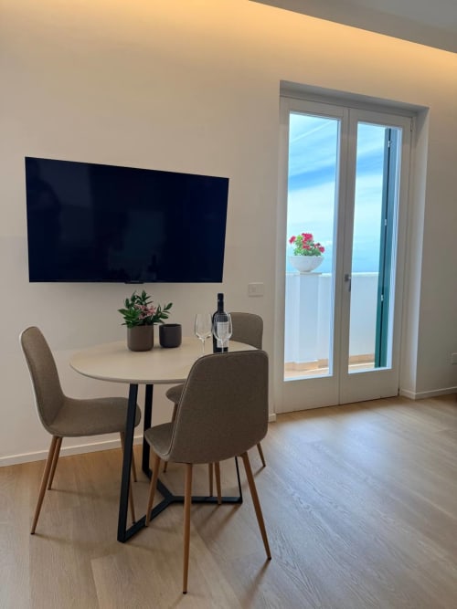 Modern dining area in Adagio Ravello's Boogie room with sea view balcony, round table, gray chairs, flat-screen TV, and