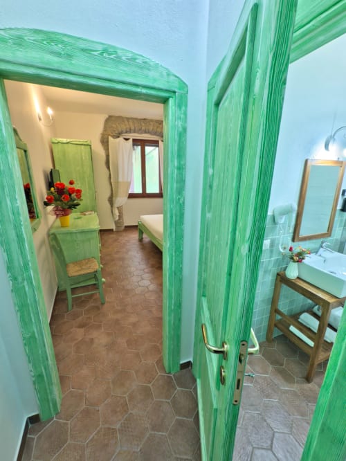 Luminose porte in legno verde che si aprono verso la camera e il bagno delle Boutique Rooms Regnos Altos a Bosa, Italia, con piastrelle esagonali