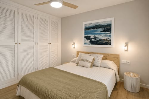 Modern bedroom with wooden headboard, white striped bedding, and coastal landscape artwork at Finques Garcia Enrich apartment
