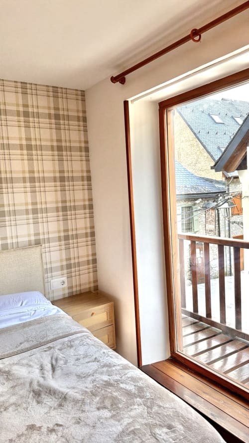 Dormitorio con cama, cortinas de cuadros, puerta-ventana de madera con vistas a los tejados de montaña en el apartamento de Vielha
