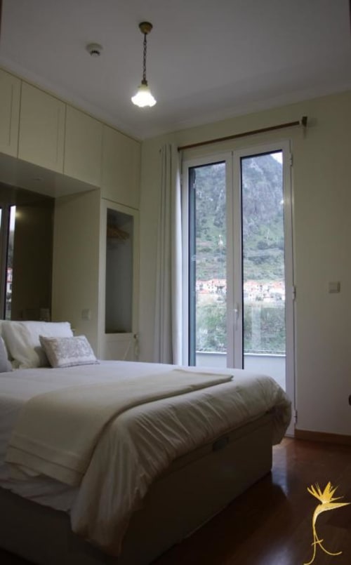 Quarto luminoso na Casa Estrelícia com cama de casal, vista para as montanhas pelas portas de vidro, móveis creme e comodidades modernas.