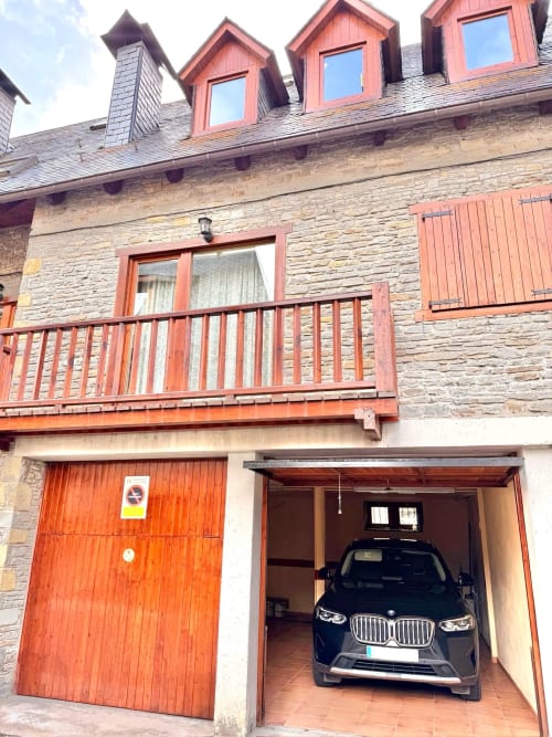 Edificio de piedra con detalles de madera roja, balcón y BMW en garaje en Eth Narjò en Vielha