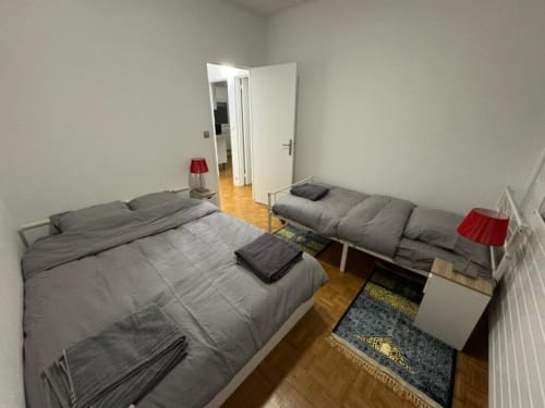 Deux lits gris dans une chambre lumineuse à Kawichimmo Locations, Louvres, avec porte blanche et lampes rouges