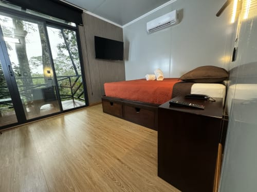 Habitación moderna del Contenedores Boutique Hotel con vista al jardín, cama con ropa de cama naranja, TV, aire acondicionado y acabados de madera