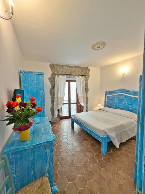 Camera luminosa a Regnos Altos a Bosa con mobili blu in legno, letto matrimoniale, fiori e accesso al balcone