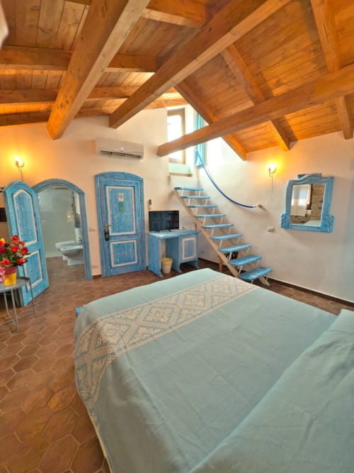 Spaziosa camera da letto con travi in legno a vista, arredamenti blu e pavimento in cotto a Regnos Altos Boutique Rooms a Bosa