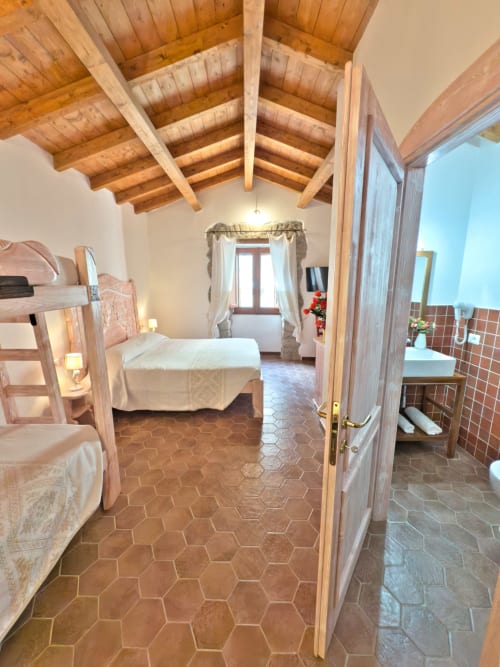 Ampia camera da letto a Regnos Altos Bosa con travi in legno a vista, piastrelle in terracotta, letti gemelli e bagno privato