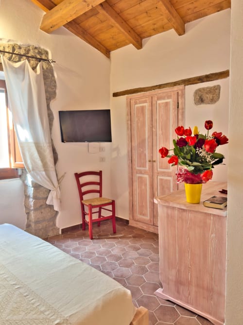 Camera rustica a Regnos Altos Boutique Rooms a Bosa con travi in legno, sedia rossa, armadio rosa e fiori rossi freschi