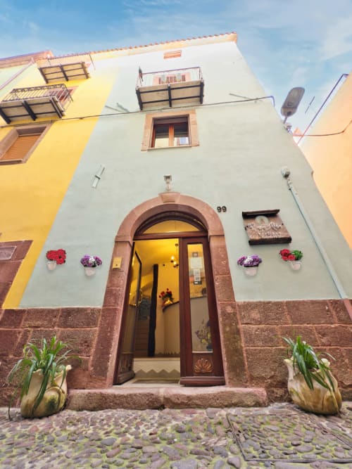 Incantevole ingresso di Regnos Altos Boutique Rooms a Bosa, con porta in legno ad arco e fiori colorati