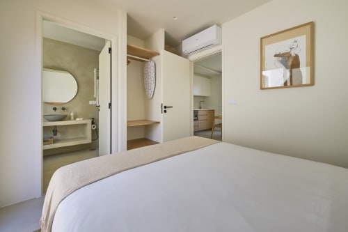 Quarto moderno com casa de banho privativa em Casa Redonda, Aroeira. Cama branca, roupeiro em madeira e entrada espelhada para a casa de banho.