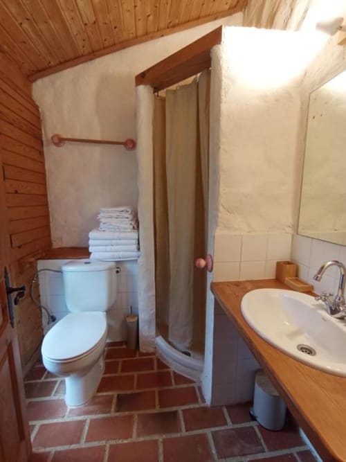 Baño de Casa Aldea L'Ayalga con inodoro, lavamanos, ducha, toallas blancas y piso de barro rústico