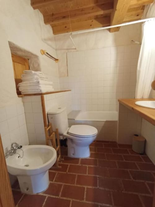 Baño espacioso en Casa Aldea L'Ayalga con doble lavamanos, inodoro, ducha, estantería de madera y piso de baldosas de terracota