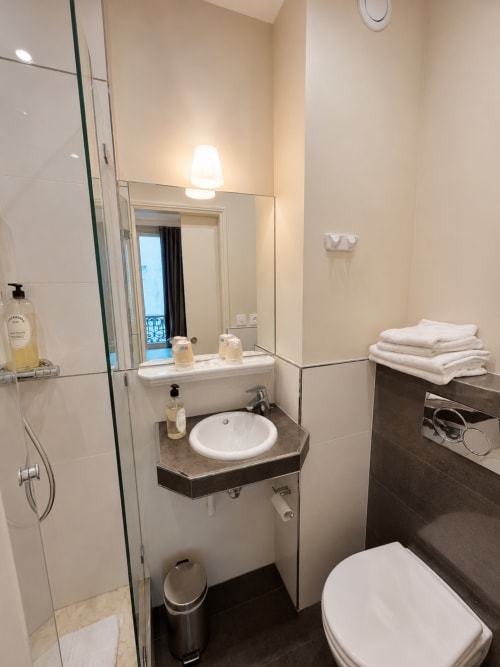 Salle de bains moderne de l'Hôtel des Bains avec lavabo rond, toilettes, douche et serviettes blanches sur étagères sombres