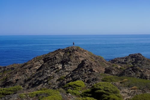 Solitary hiker stands atop rocky coastal peak overlooking Mediterranean Sea near Hotel de la Font Perelada, Girona