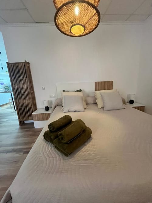 Dormitorio moderno en Casa Portalet de Xátiva con cama blanca, toallas de oliva, lámpara de techo tejida y muebles de madera