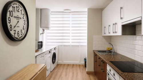 Cocina moderna con encimera de granito, armarios blancos, lavadora y reloj de Kensington Station en apartamento de Baiona