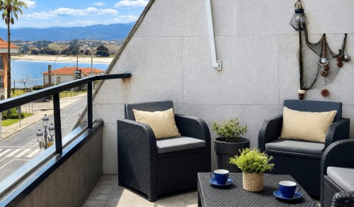 Terraza con vistas al mar de Baiona con sillas de mimbre negro, cojines amarillos y océano de fondo en el apartamento Holibai Luceira