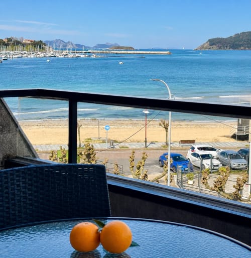 Balcón con vistas al mar de apartamento en primera línea de playa de Baiona, naranjas sobre la mesa, puerto deportivo y playa de arena abajo