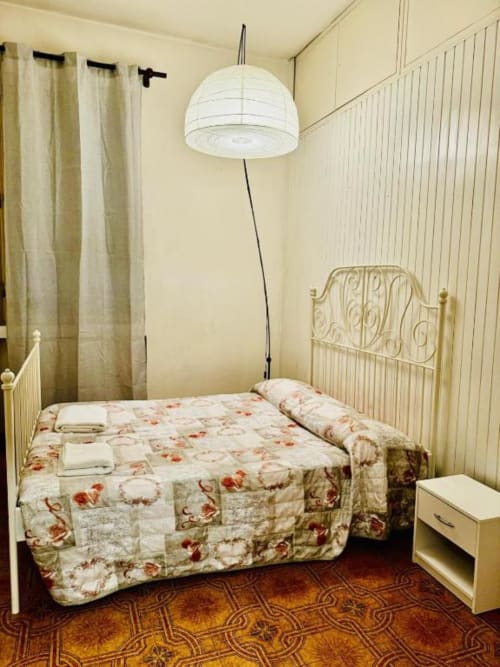 Camera accogliente di BnB Fantasia a Bologna con biancheria floreale, letto in ferro, lampada a sospensione bianca e tende color crema