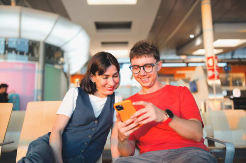 Joven pareja sonriente mirando un smartphone naranja en el moderno vestíbulo del hotel Barrosa Home, España