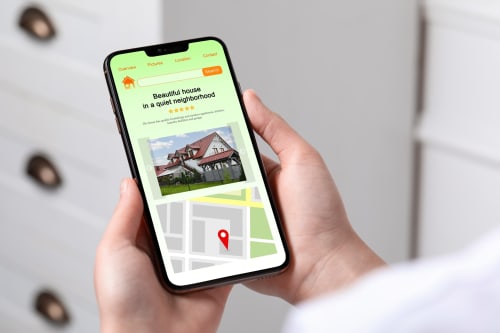 Mano sosteniendo smartphone mostrando anuncio de Barrosa Home con foto de la vivienda, calificación de estrellas y ubicación en mapa de España