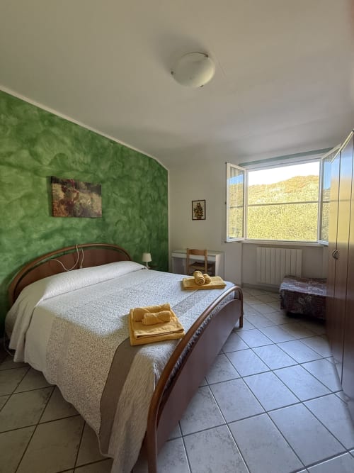 Camera luminosa con parete verde salvia, letto in legno con lenzuola bianche, asciugamani gialli, piastrelle in terracotta e vista sulla campagna