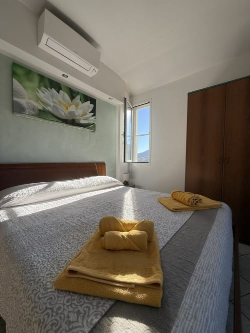 Bright bedroom at Agriturismo Gli Ulivi with queen bed, yellow towels, mountain view window, AC unit, and white lily artwork.