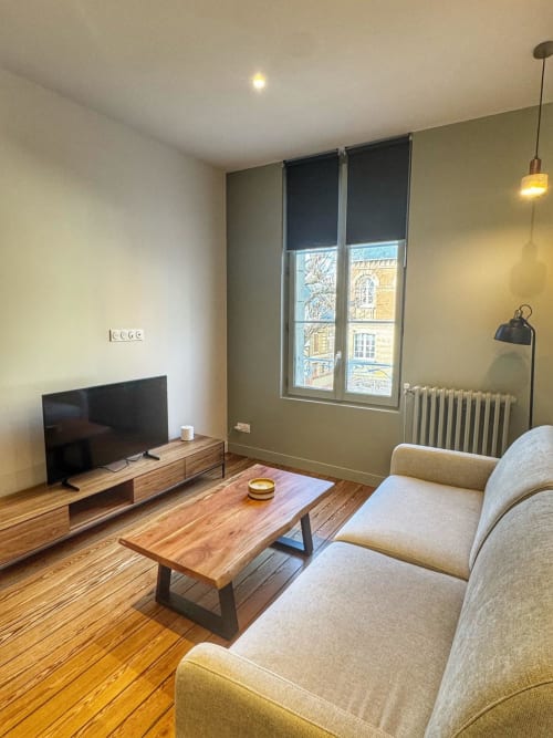 Salon moderne d'un studio à Trouville-sur-Mer avec téléviseur, mobilier en bois, canapé beige et vue sur la ville