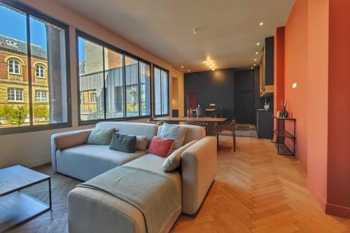 Appartement loft moderne à O LIT Trouville-sur-Mer avec canapé gris, mur d'accent orange, parquets et grandes fenêtres