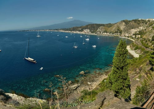 Barche a vela ancorate nelle acque cristalline del Mediterraneo vicino l'Etna, villaggio costiero siciliano con collina verdeggiante