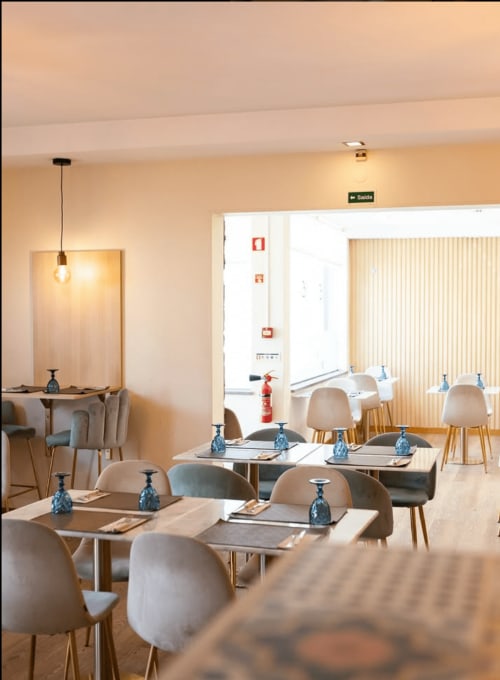 Modern restaurant interior at Arrabia Guest Houses with beige chairs, wooden tables, pendant lighting, and exit sign in