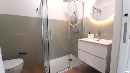 Bagno moderno nella camera d'hotel a Bari con doccia in vetro, lavabo bianco, specchio rotondo e piastrelle grigie