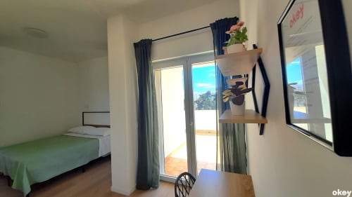 Camera da letto luminosa con biancheria verde, scrivania in legno, mensole con piante e porte in vetro che si aprono sulla terrazza con vista giardino
