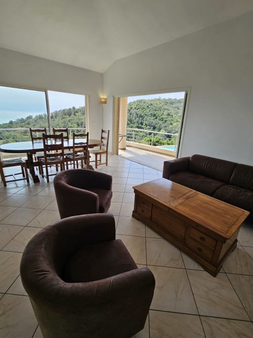 Salon de la Villa Eva avec vue sur mer, fauteuils en cuir noir, table en bois et vue forestière depuis le balcon géré par la conciergerie Corsu Di Core