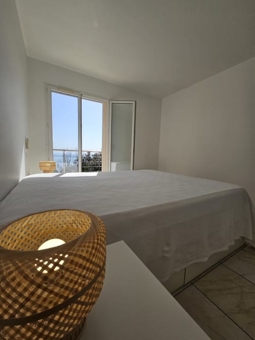 Chambre lumineuse à la Villa Eva gérée par la conciergerie Corsu Di Core avec balcon vue mer, lit blanc et lampadaire en rotin à Solenzara