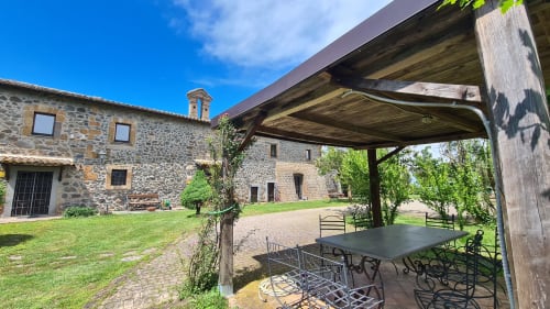 Cortile dell'Agriturismo Buriano con edifici in pietra rustica, pergolato in legno, tavolo da pranzo e lussureggiante giardino a Lubriano, Italia