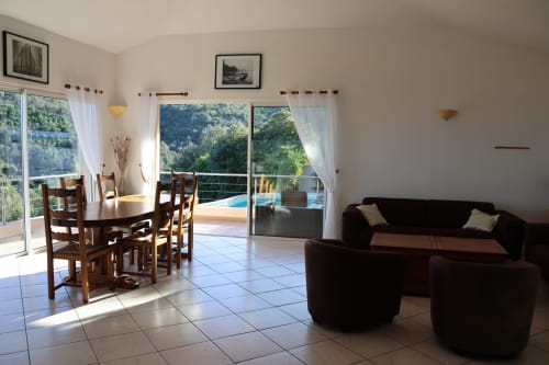 Salon de Villa Eva avec coin repas, canapé noir, terrasse vue sur la piscine et paysage corse à Solenzara