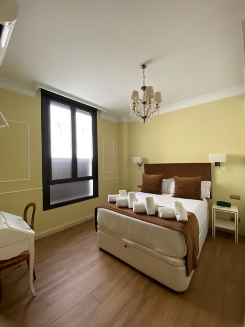 Elegant hotel suite bedroom in Seville with yellow walls, brown bed, chandelier, and city view windows