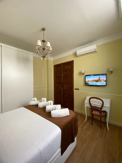 Modern hotel bedroom suite in Seville with yellow walls, queen bed, flat-screen TV, chandelier, and dark wooden doors
