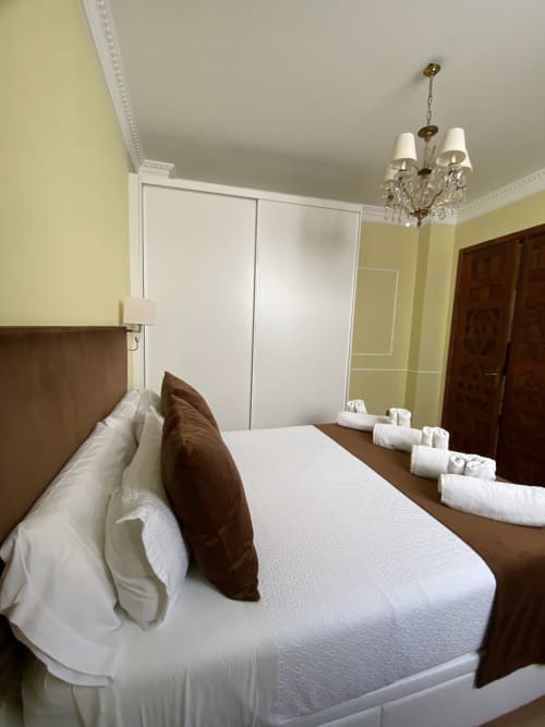 Modern bedroom in Seville hotel suite with white bedding, brown pillows, white wardrobe, chandelier, and rolled towels on bed