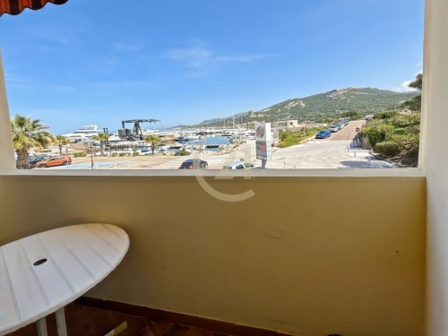 Vue depuis un balcon de la marina de L'Île-Rousse avec voiliers, plage et collines boisées sous un ciel bleu