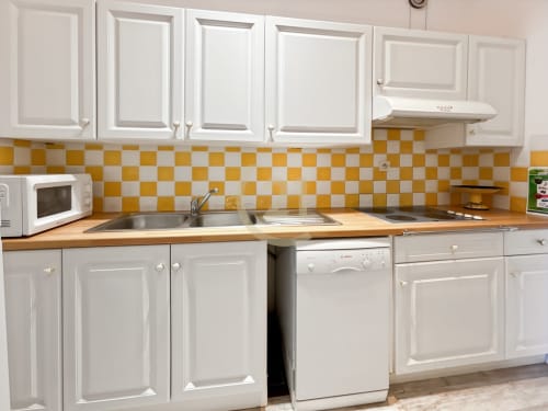 Cuisine lumineuse avec armoires blanches, plan de travail en bois, dosseret à carreaux jaunes et blancs, lave-vaisselle et micro-ondes