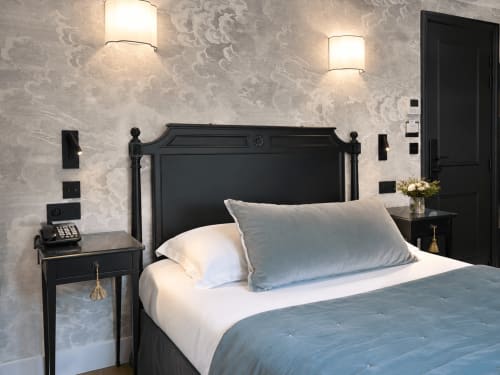 Chambre élégante de l'Hôtel des Bains avec lit en fer forgé noir, draps blancs, papier peint motifs et appliques murales