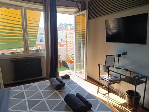 Soleggiata camera d'hotel a Roma con vista sulla città, TV, area lavoro, tappetini yoga arrotolati su tappeto geometrico, accesso balcone al Trastevere Noir